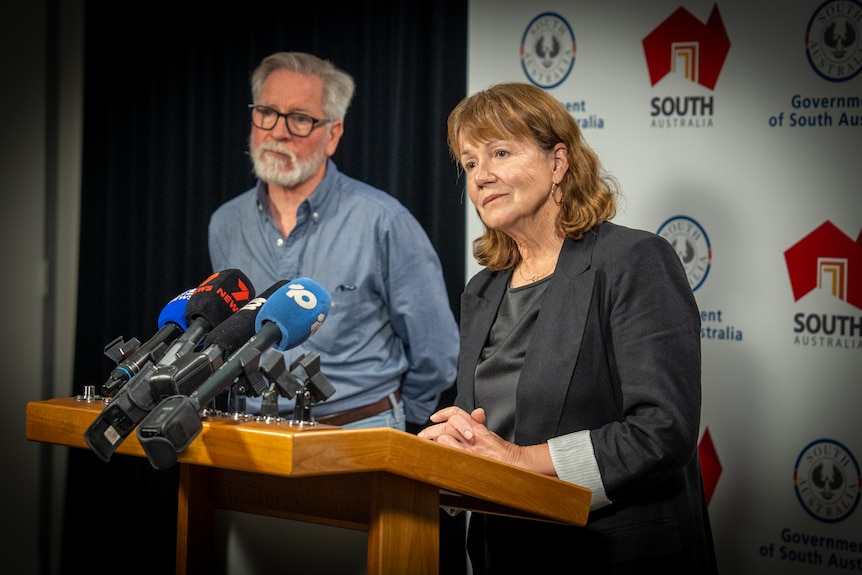 A bearded man stands near a lectern upon which a woman is resting her hands.
