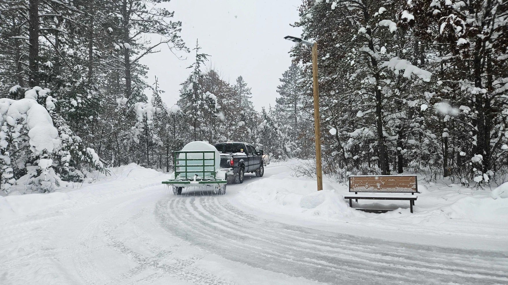 A pickup truck towing a small green trailer drives on a snowy road through a forest, passing a snow-covered bench.