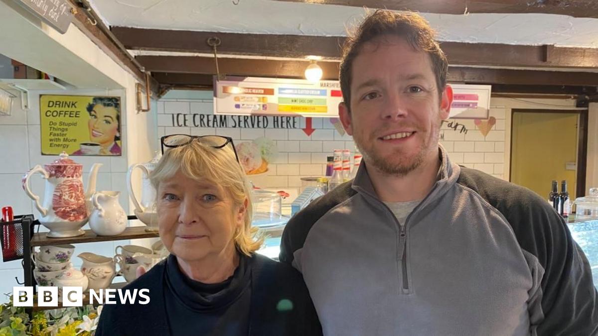 A man and a woman stand side-by-side in front of a cafe counter. The man is on the right wearing a grey quarter-zip jumper with black shoulders. The woman is in a black jumper and has her glasses resting on her head.