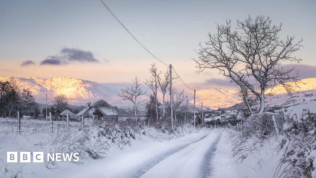Snow covered hills and a road in a rural area. Two houses with snow covered roofs are also visible