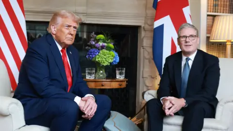 Ian Vogler/Getty Images Donald Trump and Keir Starmer sit in a formal drawing room setting  on arm chairs with US and UK  flags behind them. Both have serious expressions and are looking out and not at each other. 