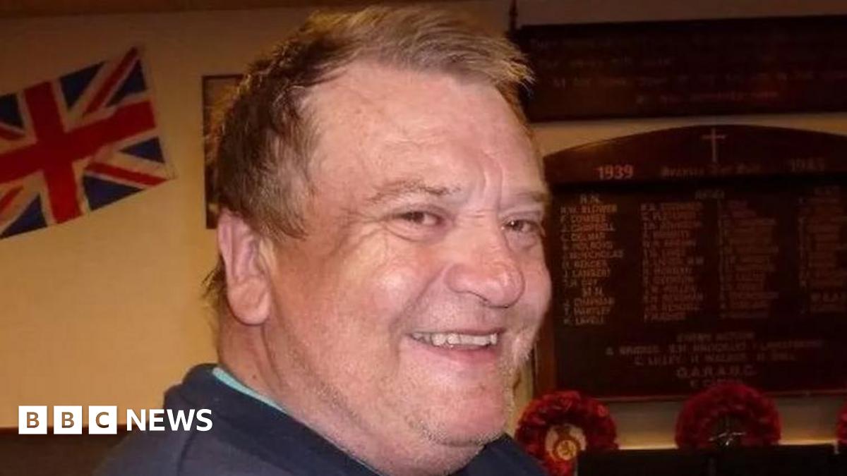 Peter Coates is looking over his shoulder and smiling brightly at the camera. He has short brown hair and is wearing a blue jumper. There is a wooden memorial board on the wall behind him, as well as a union jack flag.