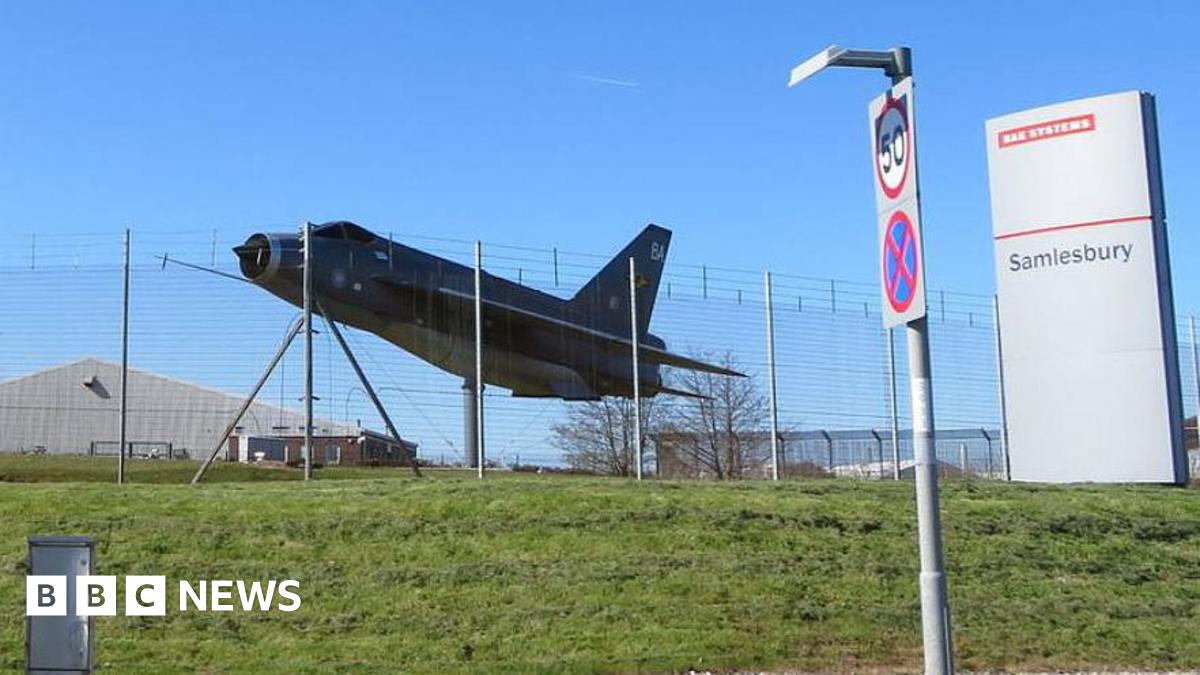 A replica of a Lightning fighter jet stands outside the main gate of the BAE Systems manufacturing site at Samlesbury, near Preston. The site is surrounded by fencing with barbed wire at the top. Outside, on a grassy bank, is a grey, vertical, rectangular block sign saying BAE Systems and Samlesbury.