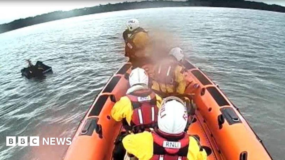 An orange lifeboat with four crew members wearing white helmets and red life jackets. There is a mattress on the water.