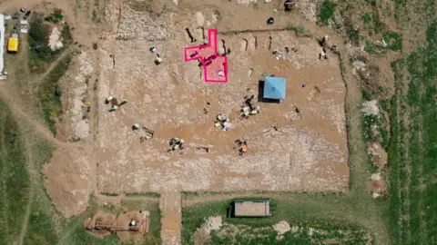 Kevin Church/BBC News Aerial view of the archaeological dig site. The location of a possible building has been graphically highlighted in pink. Six or seven graves are clustered around it.  There are another six or seven graves visible that are located further from the highlighted feature.  Archaeologists are working at different parts of the site wearing yellow high visibility jackets. The wider area of the cemetery that is being excavated has exposed brown soil. It is surrounded by a green field. 
