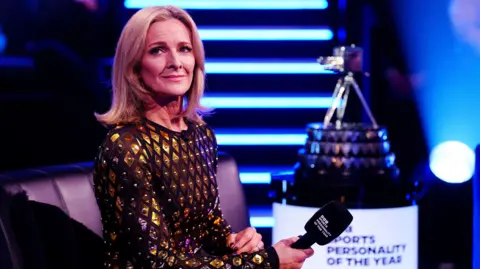 PA Media Gabby Logan wearing a black dress with gold sequence scattered across it. She is holding a microphone in her right hand and the Sports Personality of the Year award can be seen blurred in the background on her right.