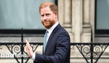 Prince Harry - a slightly-balding man with red hair and a red beard waves as he steps into a dark coloured car. He wears a pale blue shirt and a navy blue suit, with a brown beaded bracelet.