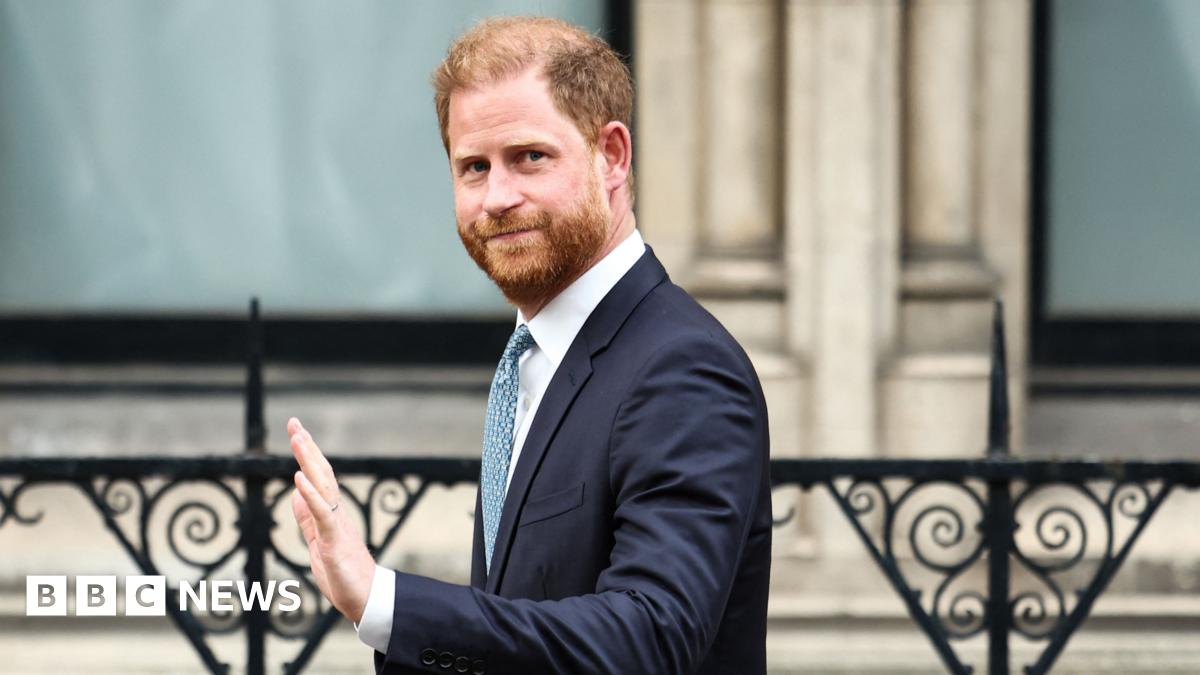 Prince Harry - a slightly-balding man with red hair and a red beard waves as he steps into a dark coloured car. He wears a pale blue shirt and a navy blue suit, with a brown beaded bracelet.