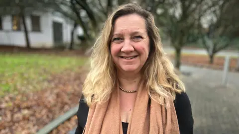 Phil Shepka/BBC Lucy Nethsingha looking into camera. She has blonde hair, and is wearing a dark jumper with a burnt orange scarf. She has trees behind her.