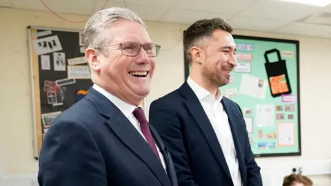BBC Sir Keir Starmer and Damian Egan stand together in a classroom where children are sitting at desks. Both me are wearing suits and the children are in school uniform. 
