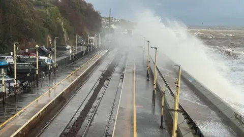 Steve Briers The picture shows huge waves crashing over a sea wall during rough weather. Brown, churning water is surging in from the right, and the force of the waves is sending spray high into the air. A railway line runs along the left side of the image, curving around the bay, with wet tracks glistening from the spray. Vehicles are parked on the left of the railway line.