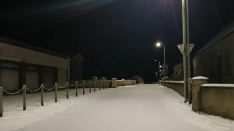 BBC NI WEATHER WATCHER - STUARTY Thick snow covers a road, the picture is taken in the dark but a street light lights up the road.