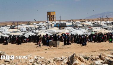 Donald Trump and Keir Starmer sit in a formal drawing room setting  on arm chairs with US and UK  flags behind them. Both have serious expressions and are looking out and not at each other.
