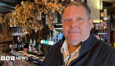 Wayne Edge stands facing the camera. He is wearing a dark jumper with a zip, and underneath a white shirt