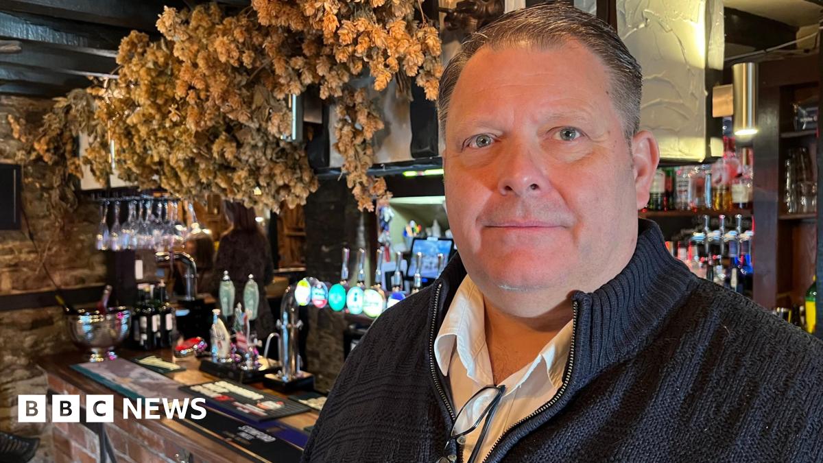 Wayne Edge stands facing the camera. He is wearing a dark jumper with a zip, and underneath a white shirt