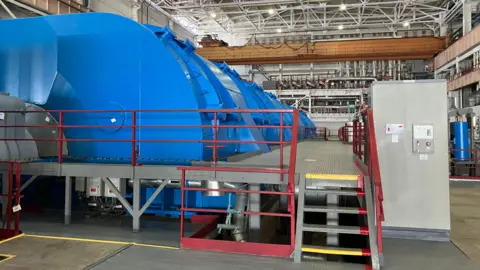The turbine hall at the nuclear power plant. A giant blue turbine sits in a large hall, with steps leading up to it. 