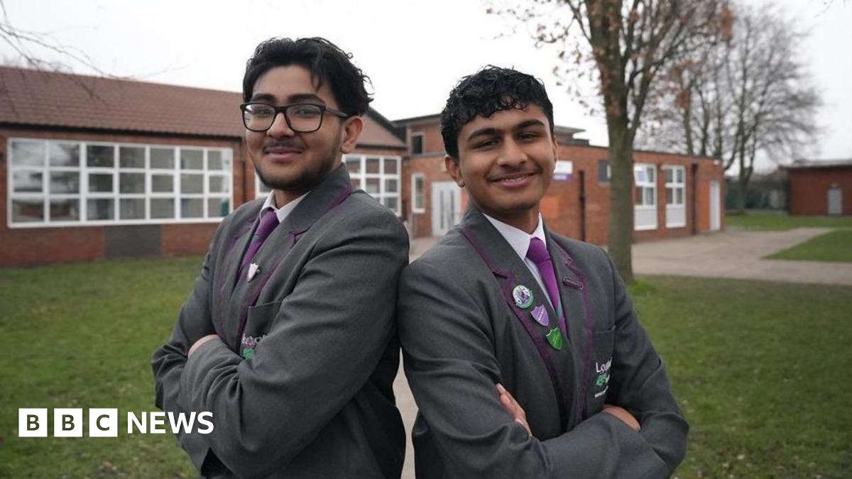 Abdullah and Bakhtiar are standing back to back with their arms folded smiling at the camera. They are wearing their gray school blazers, purple ties and white shirts.