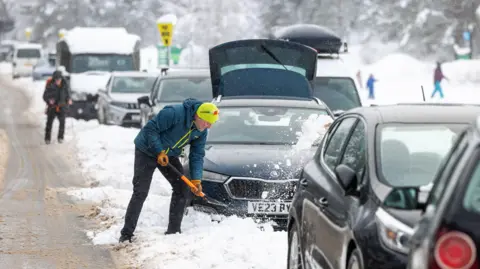 PA Media A person in winter clothing shovels snow away from a car on a congested, snow-covered road, with several vehicles stuck in traffic and heavy snowfall visible in a mountainous or rural setting.