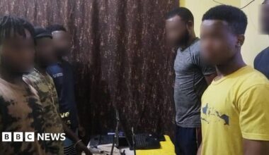 Five men stand over laptops on a table with their hands behind their backs.  The victims were found in their respective apartments, operating laptop computers.