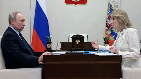 Sputnik/Mikhail Metzel/Pool via REUTERS Vladimir Putin and Maria Lvova-Belova sit on opposite sides of a desk. He is on the left, looking at her as she speaks. He is wearing a dark suit and tie, she is wearing a white dress. There is a Russian flag in the background. 