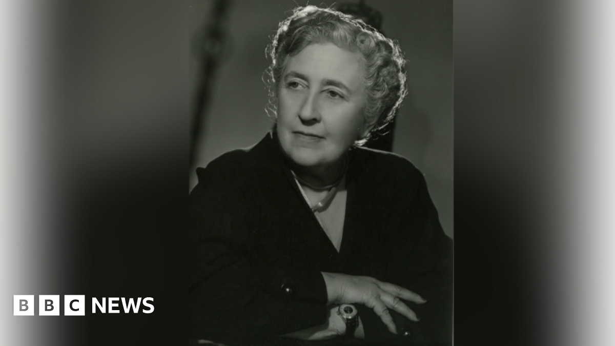 An archive sepia image of Agatha Christie, around 1950. She is sitting down with her arms folded and looking away from the camera.