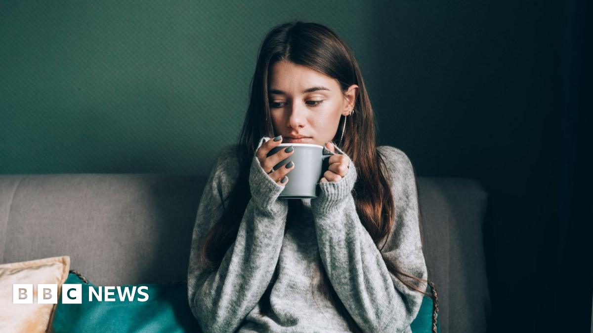 A young woman with a grey jumper on sits on a sofa and is holding a hot drink.