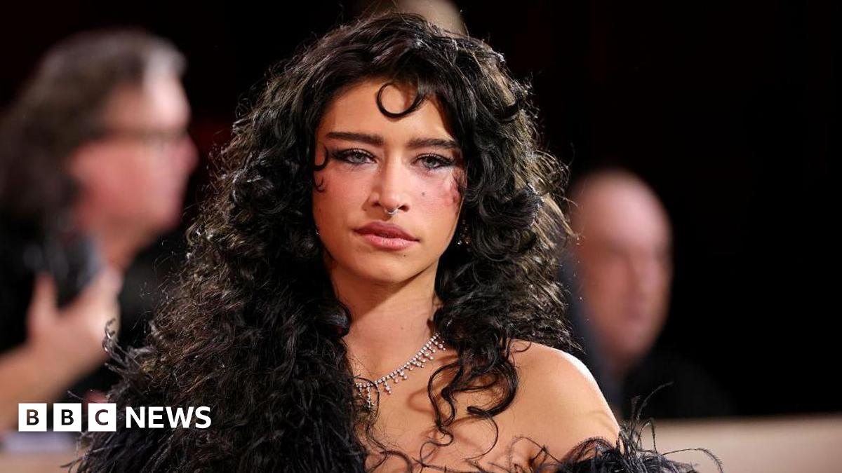 Odessa has brunette hair, she's posing for a picture on a red carpet, she has a nose ring and a silver necklace on. In the background there are blurred photographers.