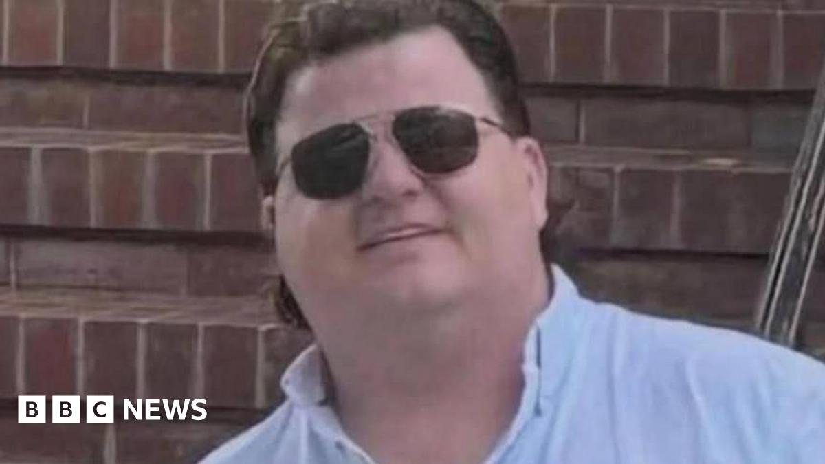 Thomas Casey has brown hair and is wearing black sunglasses and a pale blue shirt. He is smiling in front of a brick wall.