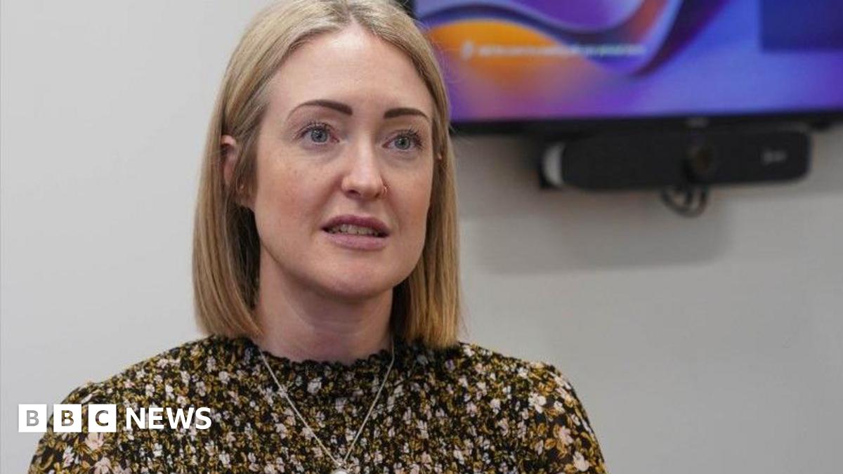 Esther Ghey, with shoulder length blonde hair and wearing a flowery top with a silver necklace, speaks to the camera