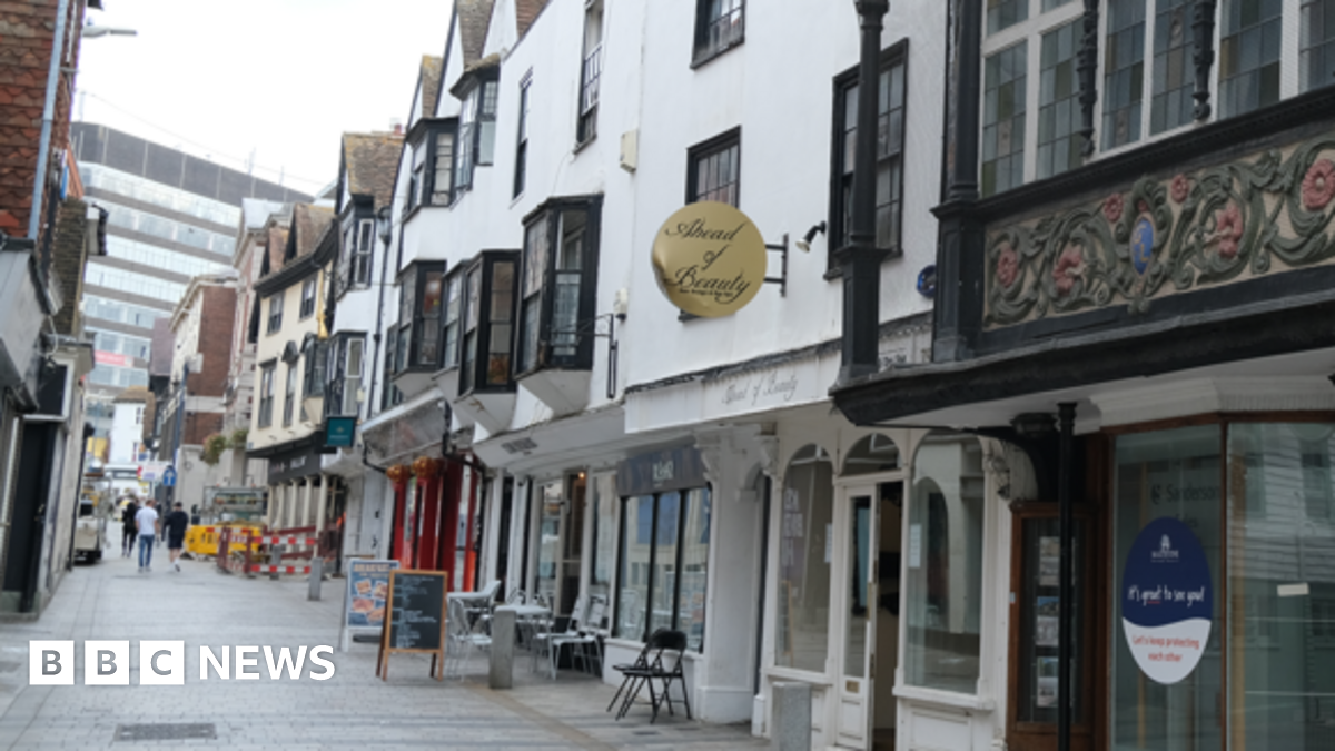 A narrow shopping street with a mix of shopfronts. Image is taken from outside on of the shops looking up the lane