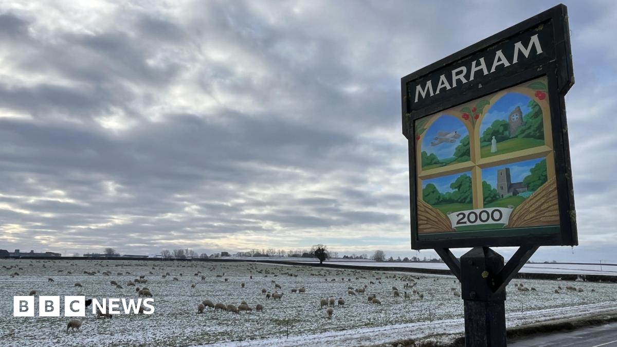 Marham village sign, with paintings of a plane, house, field and church. It stands next to a road and snowy fields, with sheep grazing.