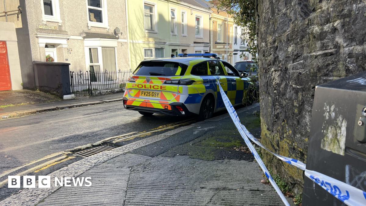 A Devon and Cornwall Police car parked at the side of a road. There is police tape from its door handle to an electricity box. The cordon is across the pavement.