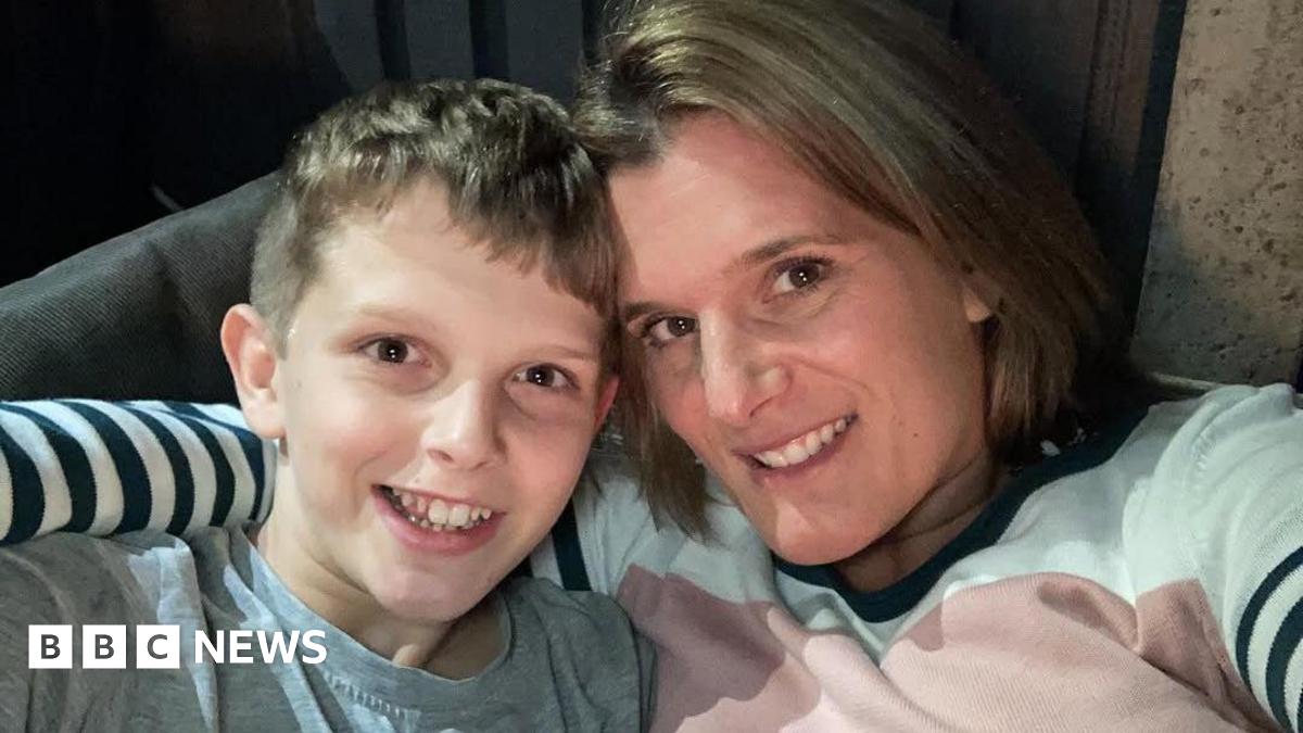 Jools (left) and Ellen (right) sit together in a chai, smiling for a selfie. Jools has short blonde hair and wears a grey top. Ellen has a blonde bob and wears a white, pink and navy striped jumper.