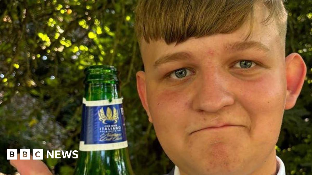 George Herd who has light brown hair cut very short at the back and in a long fringe on his forehead. He is wearing a grey jacket and a white shirt. He is holding up his right hand with an uncapped bottle and is also giving the thumbs up. Behind him are green trees and shrubs.