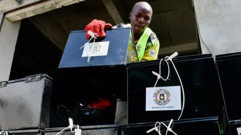 Reuters A view from below of a warehouse worker arranging black metal ballot boxes. He is wearing a hi-viz jacket and red gloves.