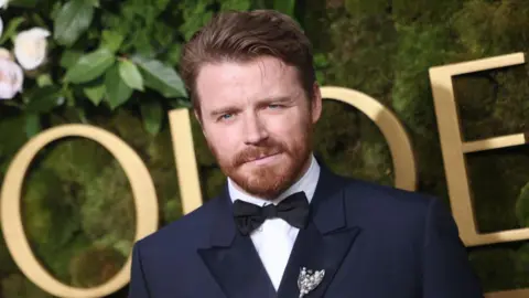 Getty Images Lowden wearing a dark formal suit with a black bow tie and a decorative brooch on the left lapel, standing in front of a green foliage backdrop with large gold letters partially visible.