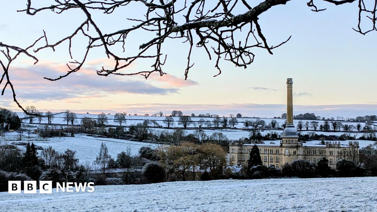 A lovely snowy scene with trees and a large building seen with a dusting of the white stuff. The sky is mostly clear with a few white and grey clouds moving away.