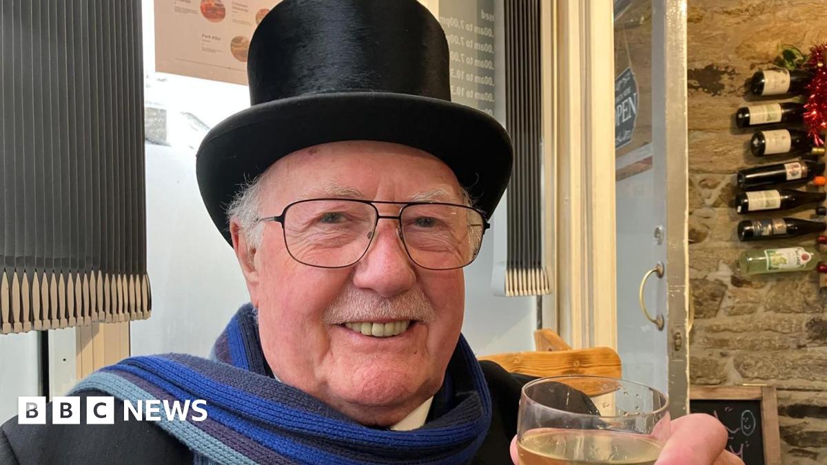 The image shows a man indoors, wearing a top hat and glasses. He is smiling at the camera and holding a glass. Behind him is a window with grey blinds, and a door.