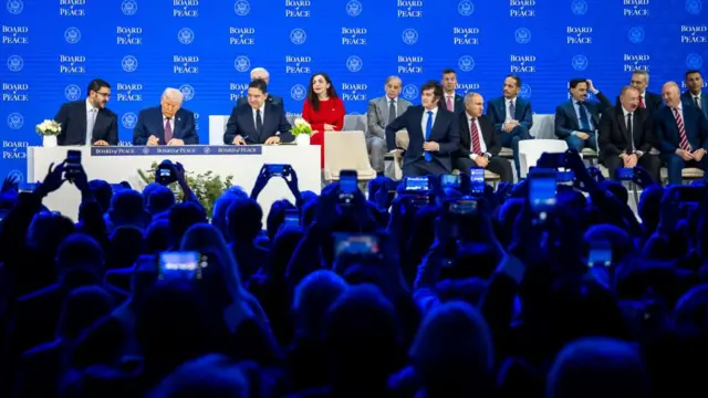 Trump signs the Board of Peace agreement as several delegates look on. An audience hold up smart phones