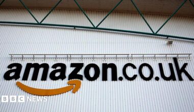 A logo sits on a wall outside the Amazon Marston Gate fulfilment centre, the building is white with green metal above.