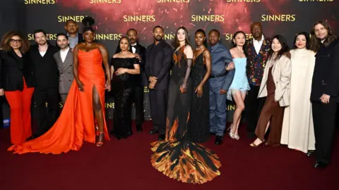 Warner Bros. Pictures Wunmi Mosaku, who is wearing an orange floor-length dress, is pictured on the red carpet for the Sinners premiere with other members of the cast and crew. 