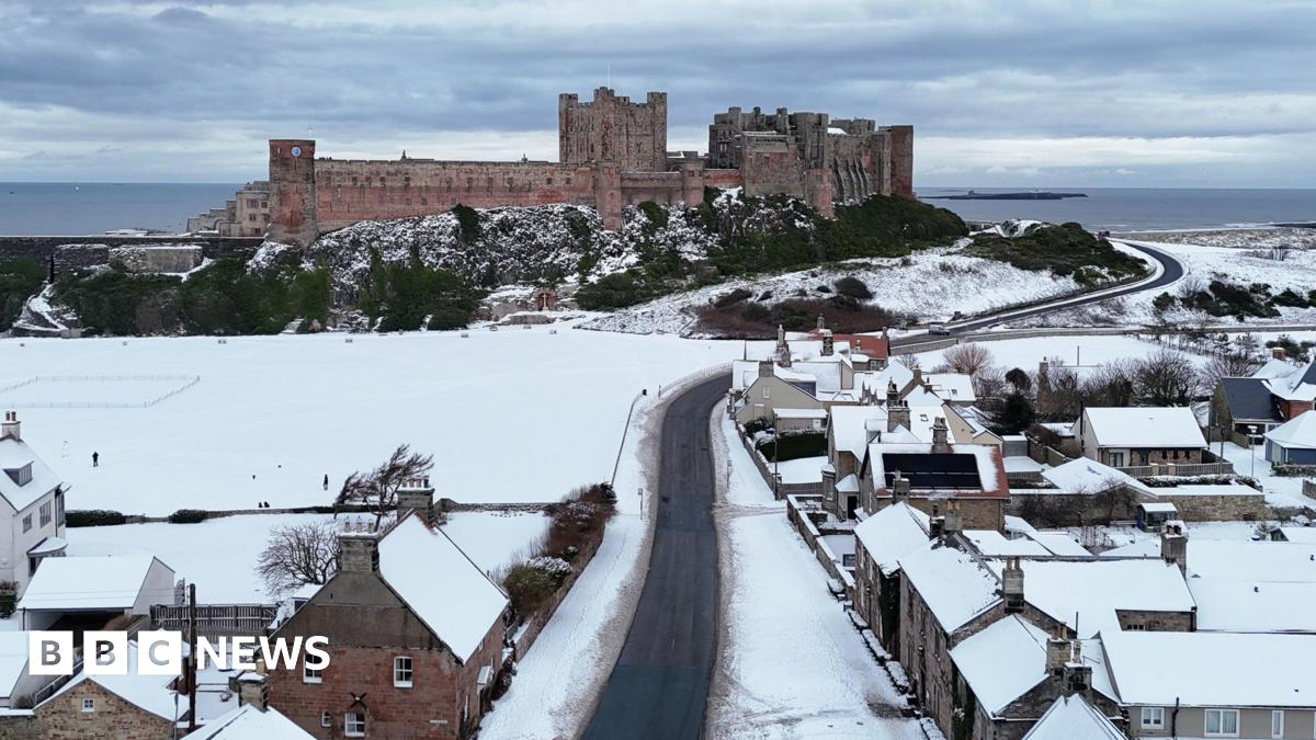 Bamburgh Castle rises above a snowy Bamburgh village. A winding narrow road leads towards the castle with the North Sea visible in the distance.