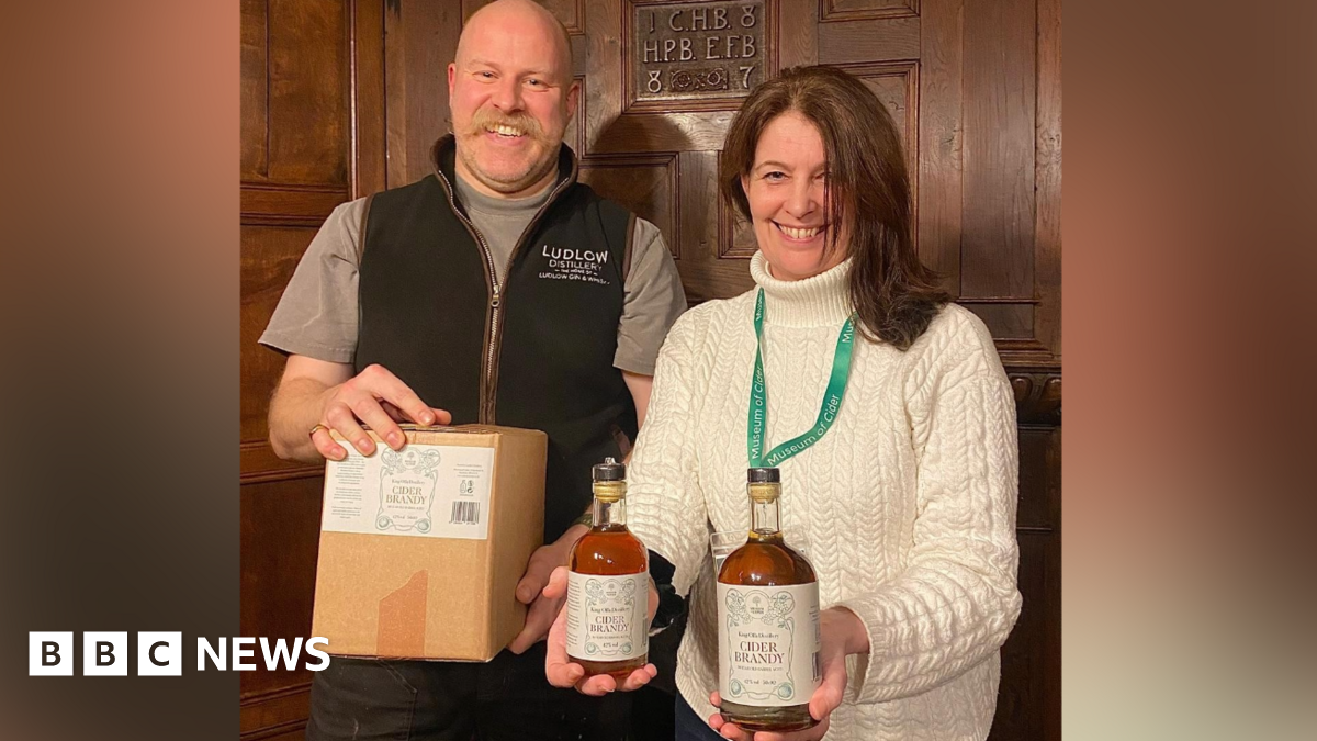 A man and a woman stand next to each other in a room with wooden panelled walls. The man is bald with a blond moustache and is holding a wooden box. The woman has long brown hair and she is holding two bottles of brown liquid.