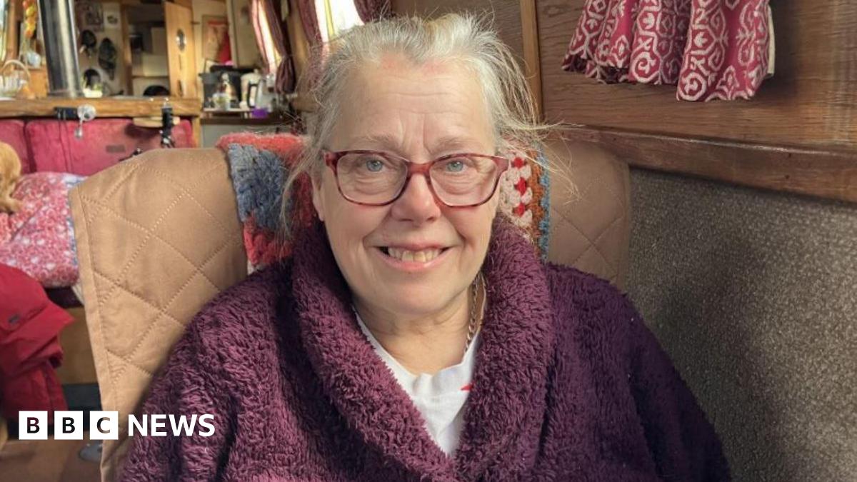 A woman with grey hair, red-framed glasses and a maroon jumper in a brown chair sitting on board her narrowboat. A small room can be seen behind her.