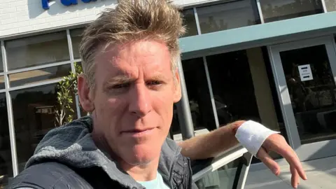 Chris Murray A man with short grey hair is resting his bandaged hand while standing in front of a hospital building.