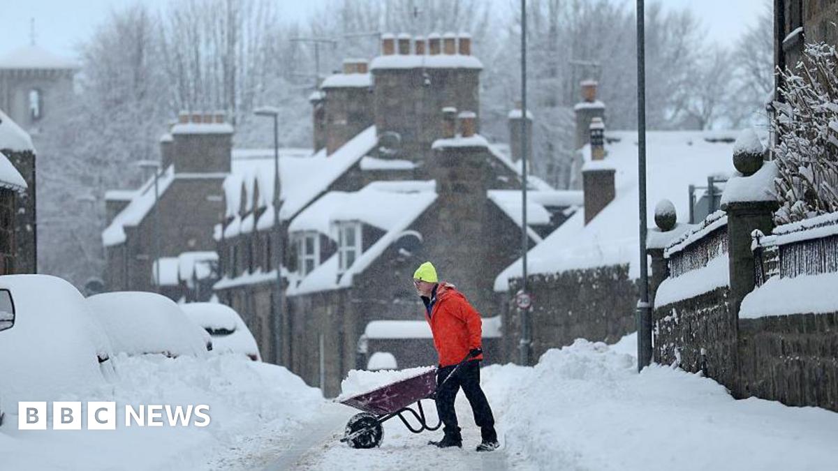 Person wearing a bright jacket and hat pushing a wheelbarrow along a snow-covered residential street lined with parked cars and stone houses during heavy winter conditions
