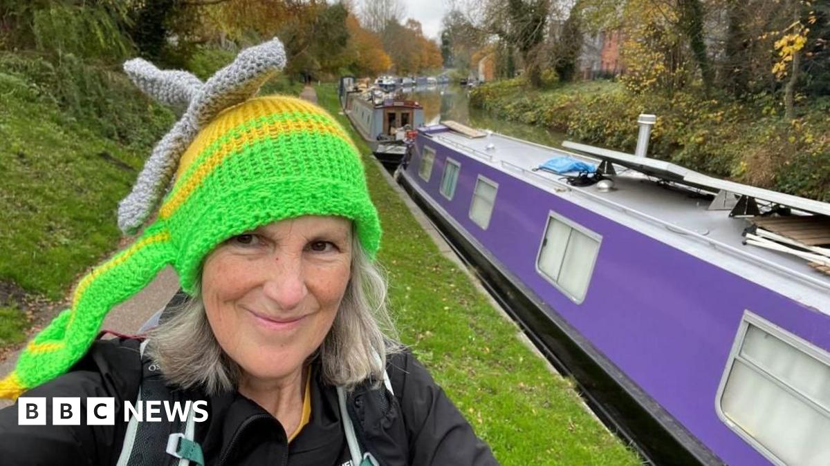 A woman with grey hair standing on a canal path wearing a knitted hat that resembles an air ambulance. A canal boat is next to her.