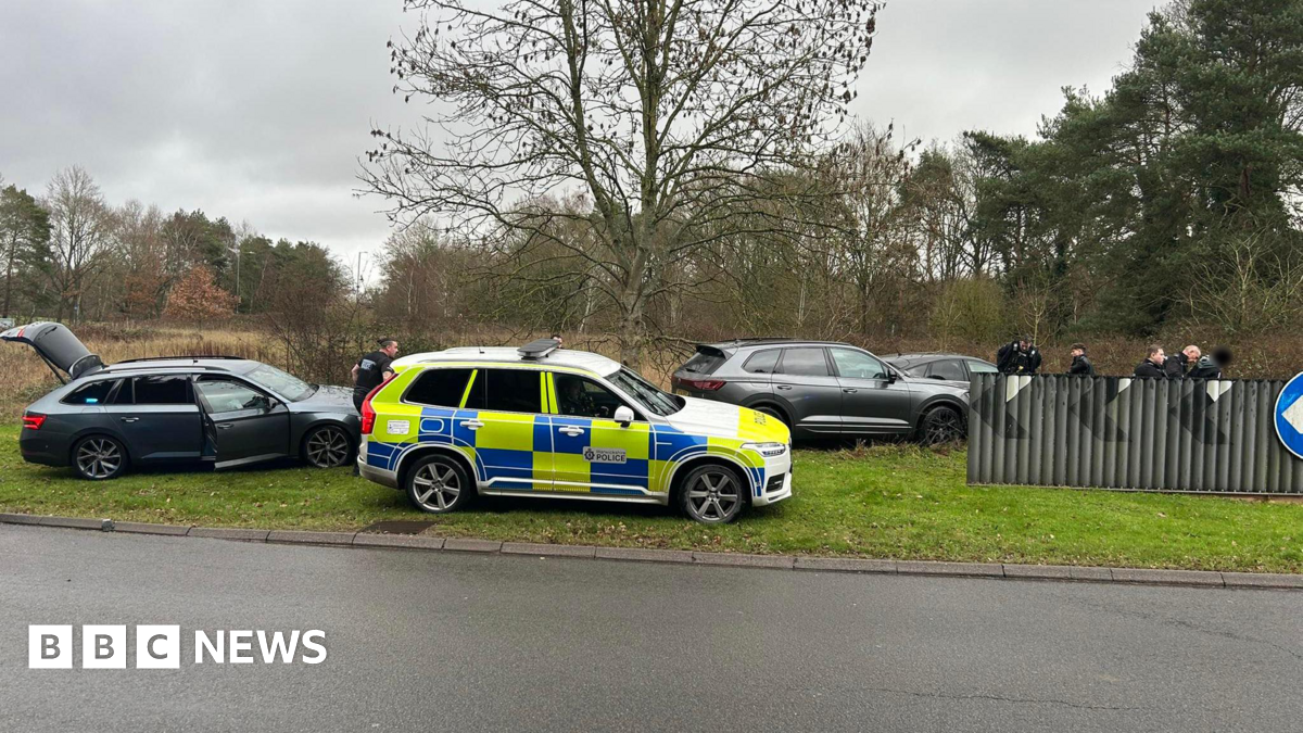 A yellow and blue police vehicle sits on a grassy bank beside the A46 in Warwickshire. Three other black vehicles are parked nearby. Police officers can be seen behind a road barrier.