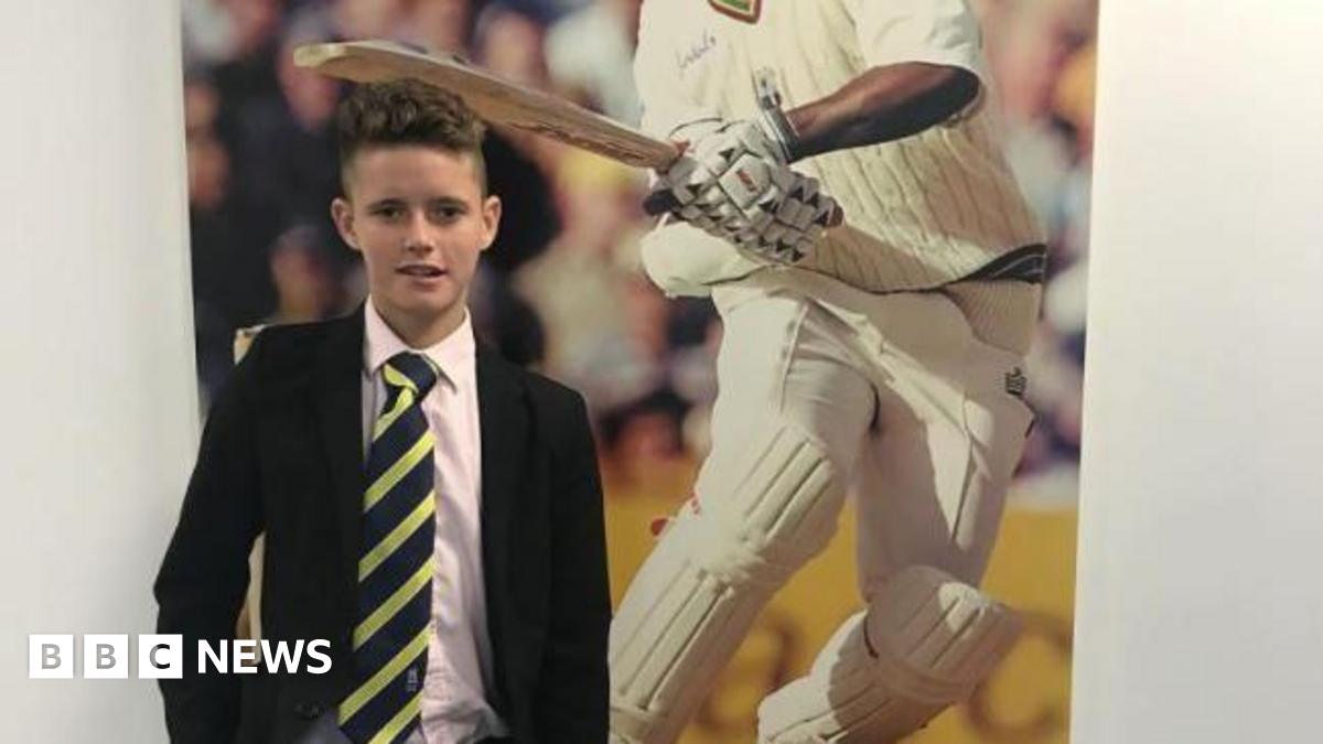 A schoolboy in a black blazer, white shirt and navy and yellow diagonally striped tie standing in front of a poster showing a cricketer.