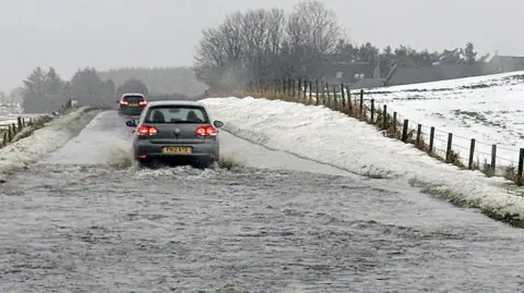 Fubar News A car drives through floodwater covering a rural road, with snow piled along the roadside and fields under wintry cond